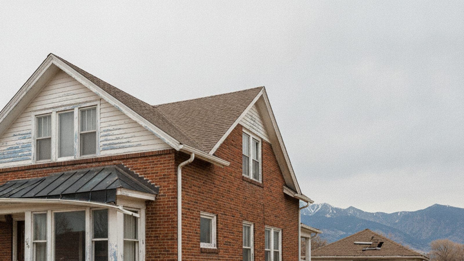 A weathered brick house with white gables and a dark bay window roof, under an overcast sky, with mountains in the background.