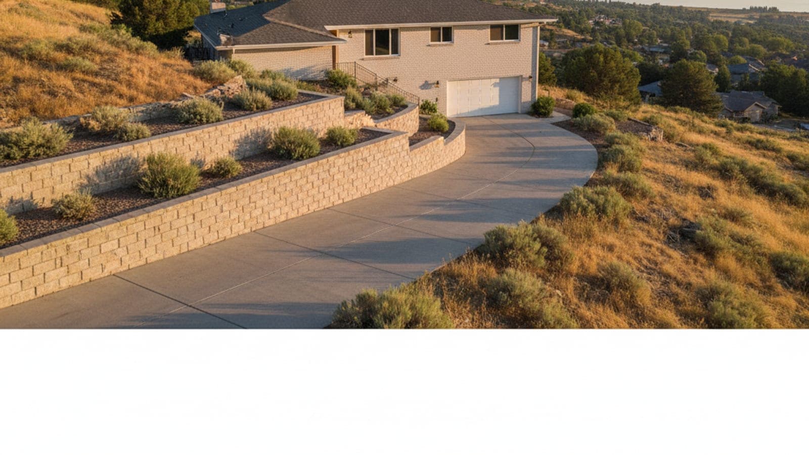A sunlit house on a hillside with a winding driveway and terraced retaining walls planted with shrubs, overlooking a valley.