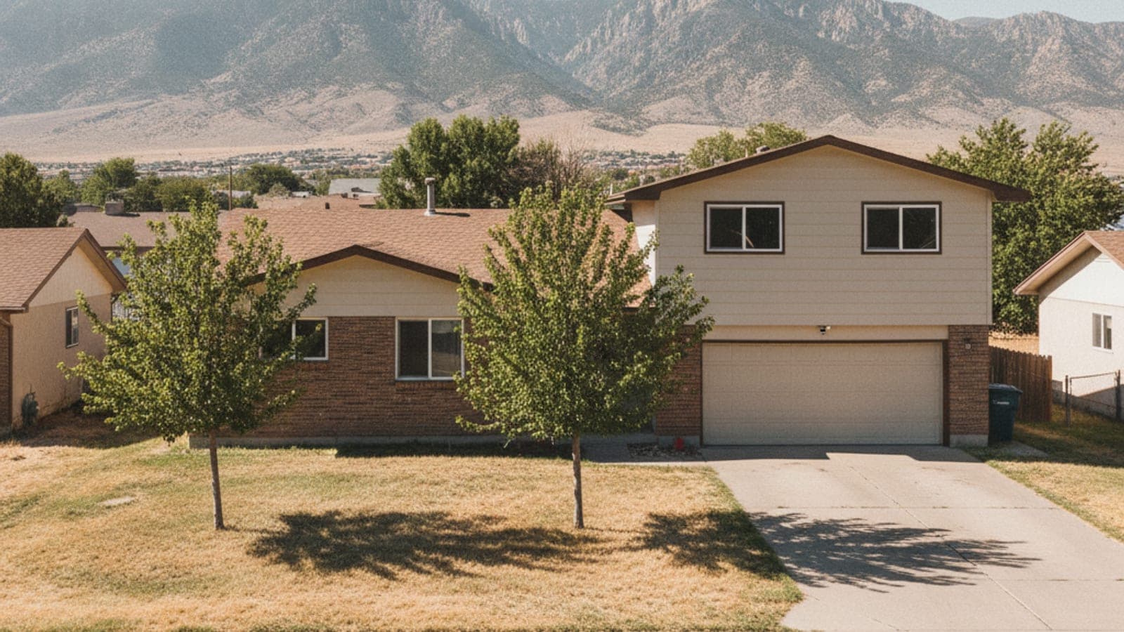 A tan house with a garage and two small trees sits on a dry lawn before large, distant mountains under a bright sky.