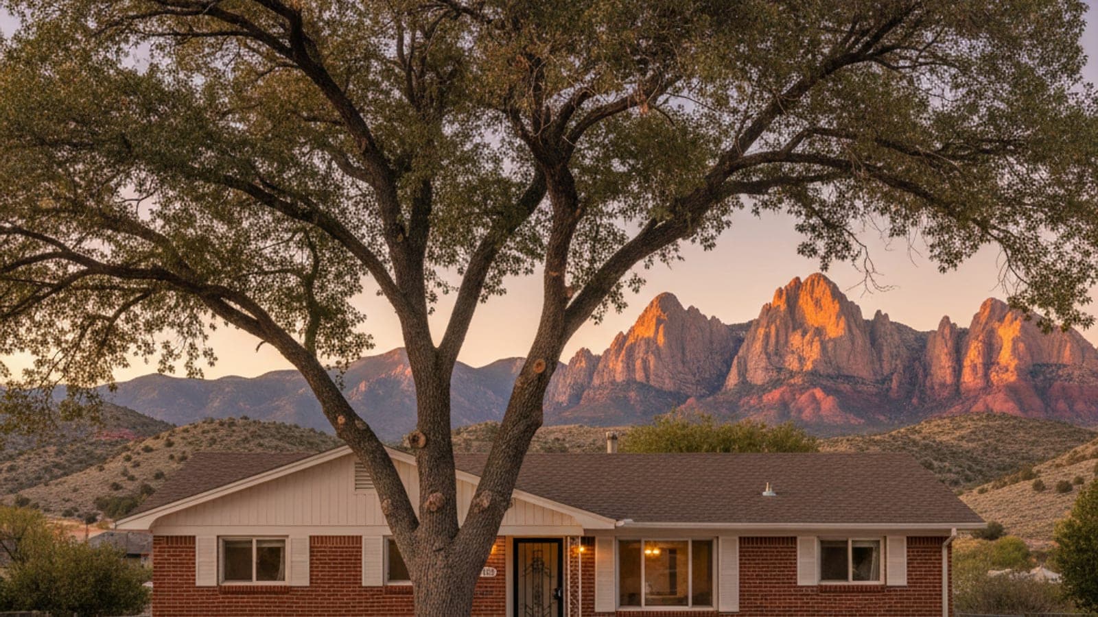 Sunlit mountains and hills rise behind a large tree and a brick house under a warm sky.