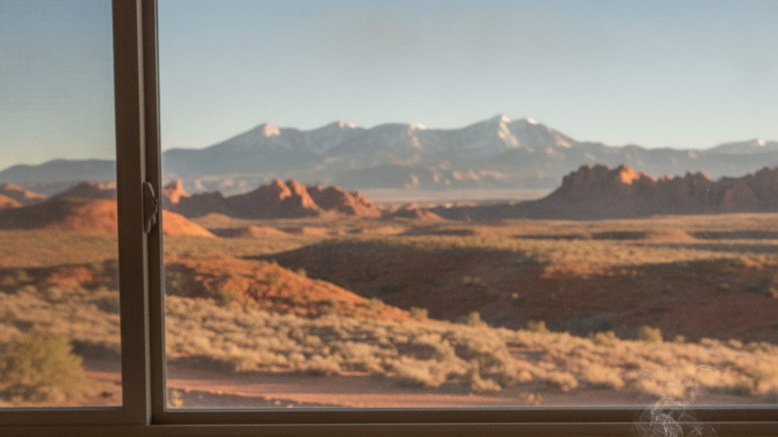 A desert landscape with red rocks and snowy mountains visible through a window frame in warm light.